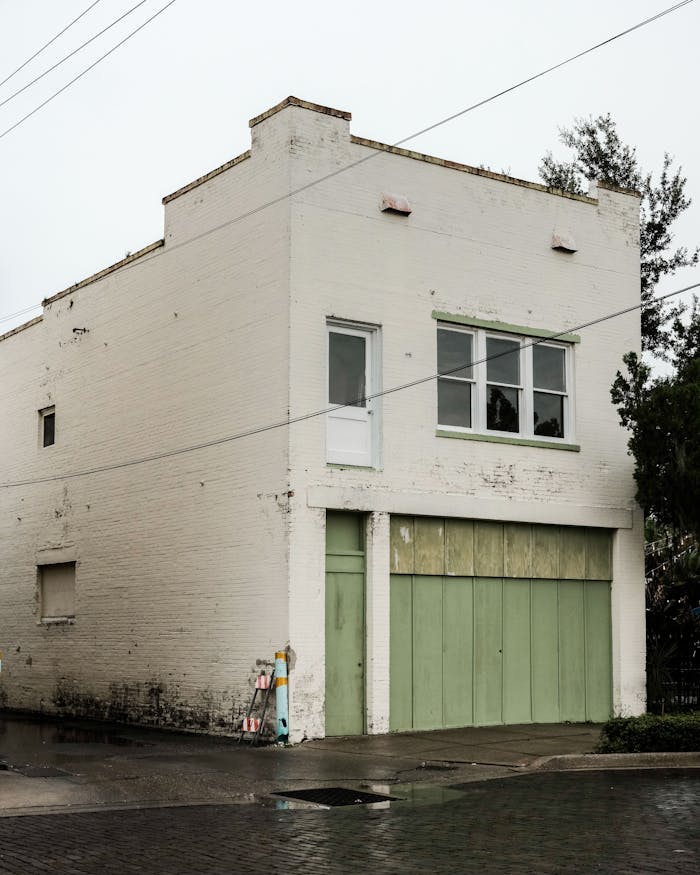 Aged white brick building with green garage doors on a rainy street corner, evokes a vintage urban feel.